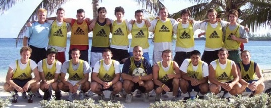 A soccer team in yellow pinnies posing together on a beach with palm trees in the background