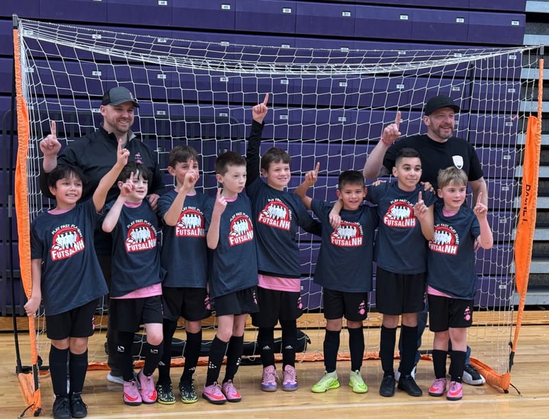 A boys team in navy FutsalNH shirts with two coaches, fingers raised, in front of a futsal goal