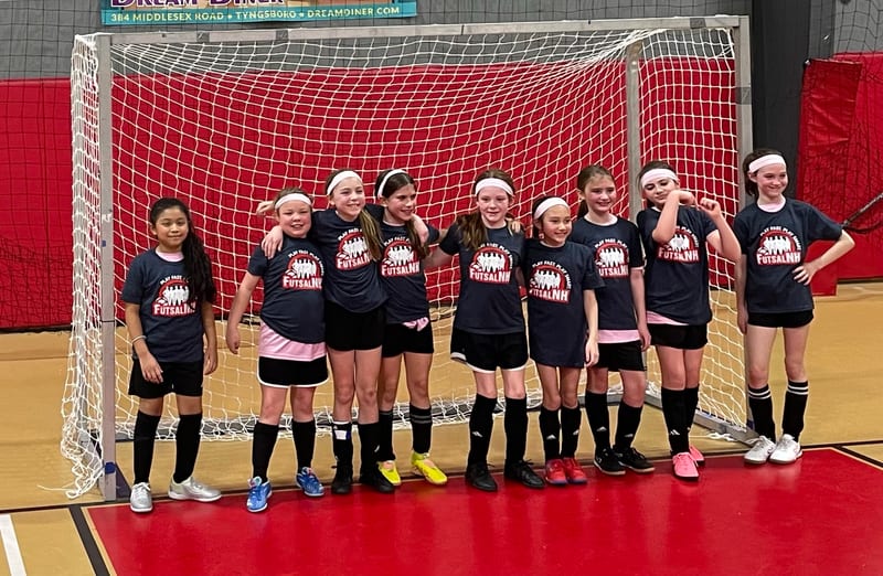 A girls team in navy FutsalNH shirts posing together in front of a futsal goal with a red backdrop