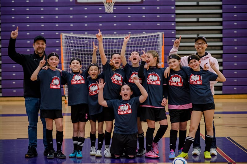 A girls team in navy FutsalNH shirts celebrating with fingers raised in front of a futsal goal