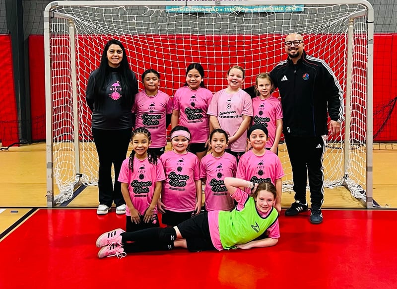 A girls team in pink Susan's Nails jerseys with two coaches posing in front of a futsal goal