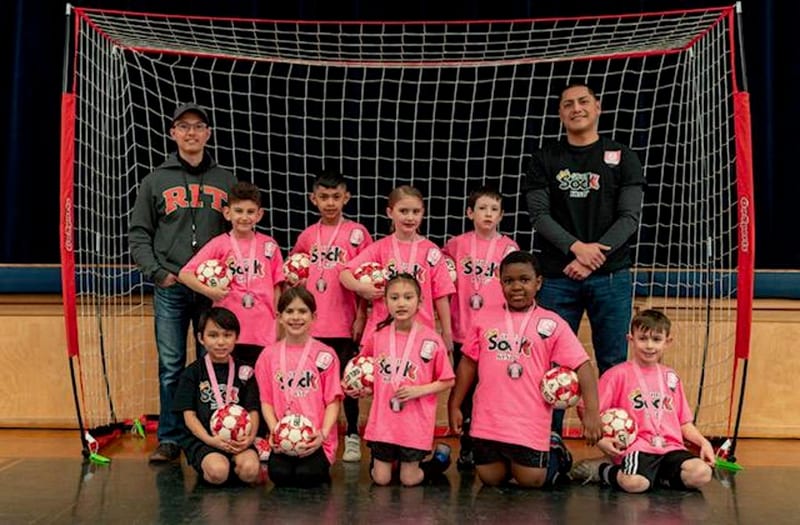 A small team in pink jerseys with two coaches posing in front of a futsal goal