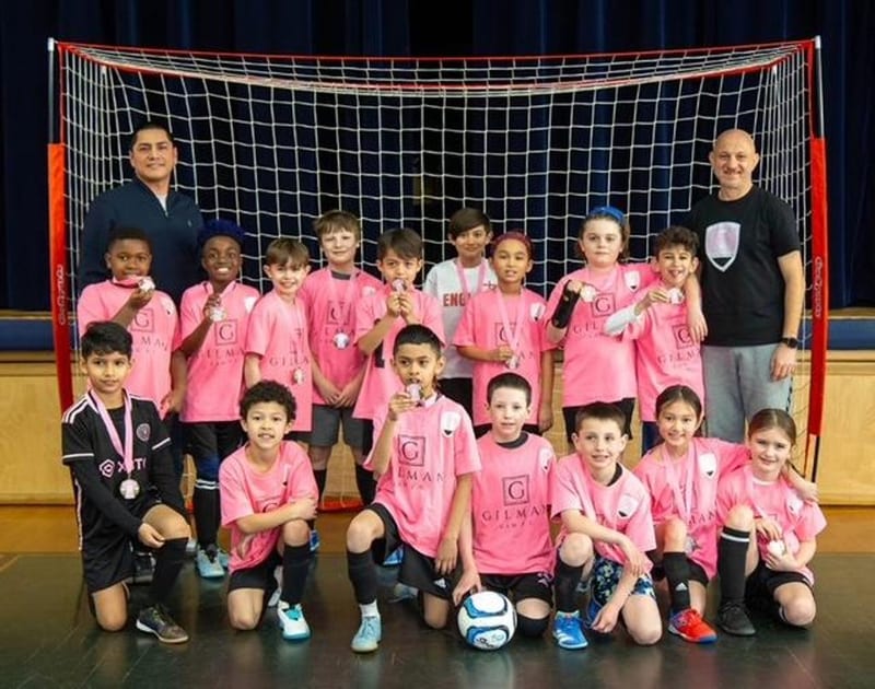 A larger team in pink Gilman jerseys with two coaches posing in front of a futsal goal, holding medals