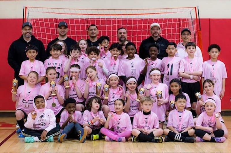 A large group of players in pink jerseys with several coaches, holding trophies in front of a red futsal goal