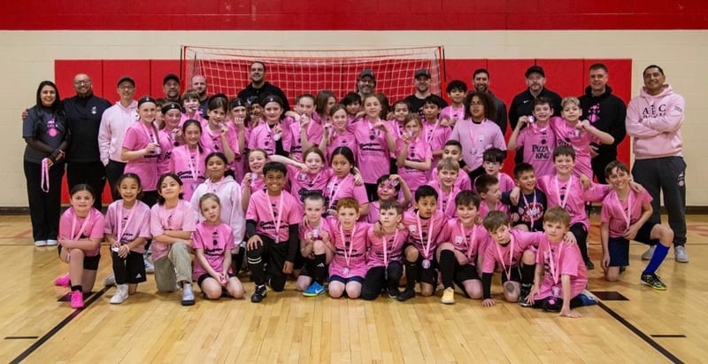 A very large group of players in pink jerseys with many coaches, wearing medals, filling a red gymnasium