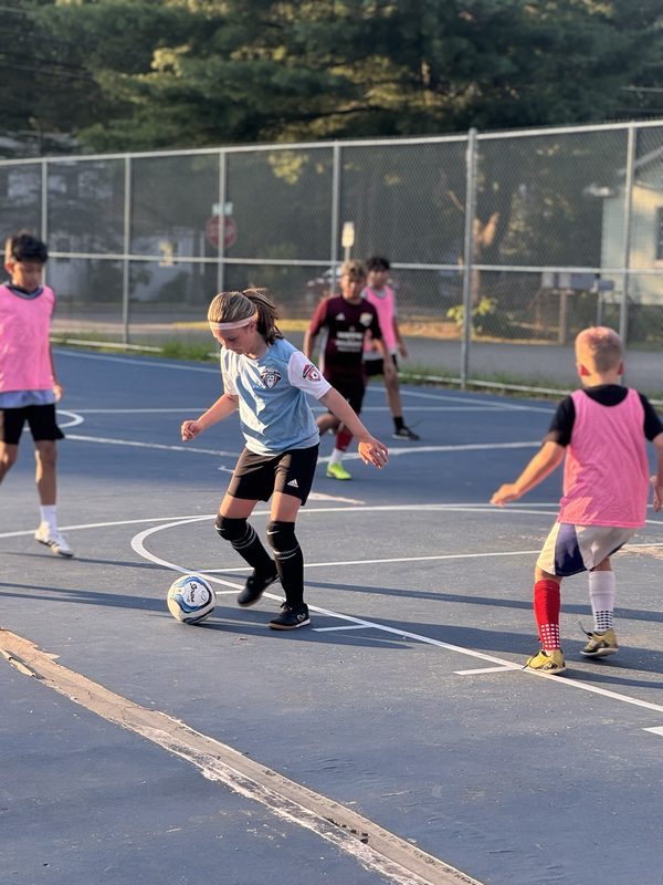 A player in a light blue jersey dribbling a ball on an outdoor court as other players close in