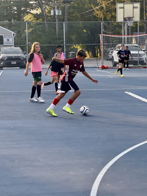 A player in a maroon jersey dribbling a ball on an outdoor court with a futsal goal and parked cars behind