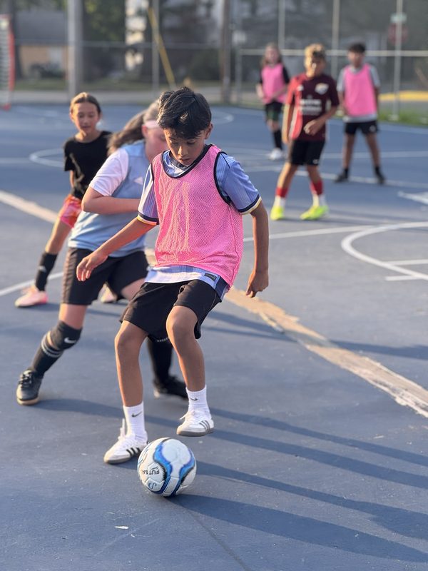 A player in a pink pinnie dribbling a ball on an outdoor court as teammates look on at dusk