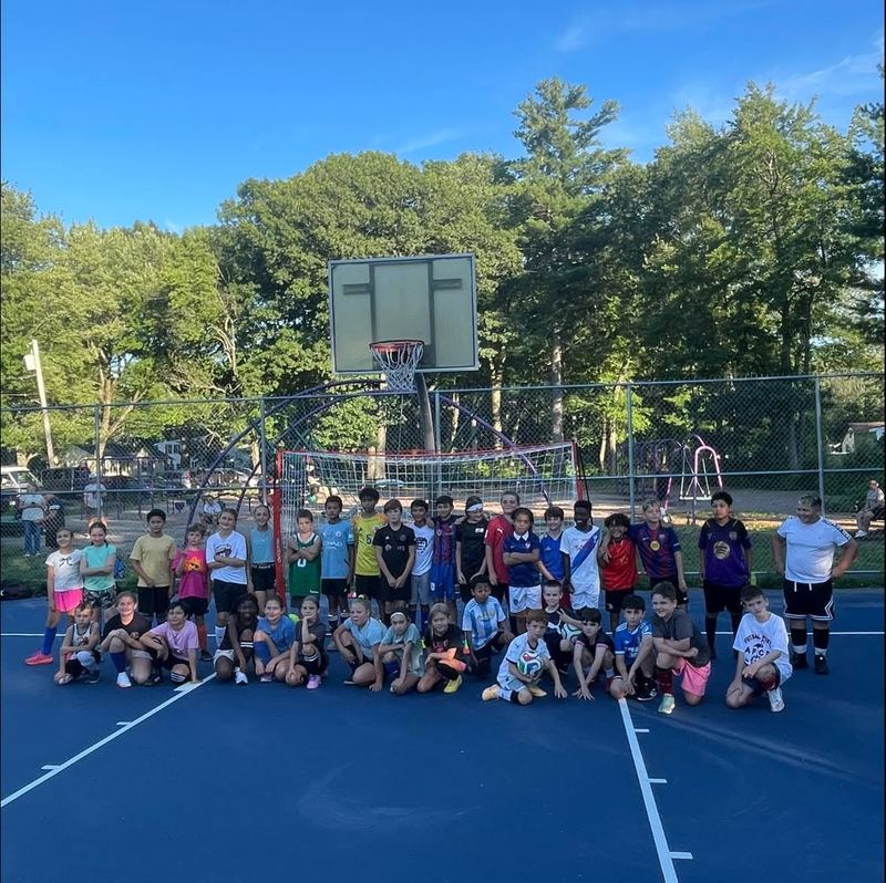 A large group of children and a coach on an outdoor blue court beside a futsal goal at dusk