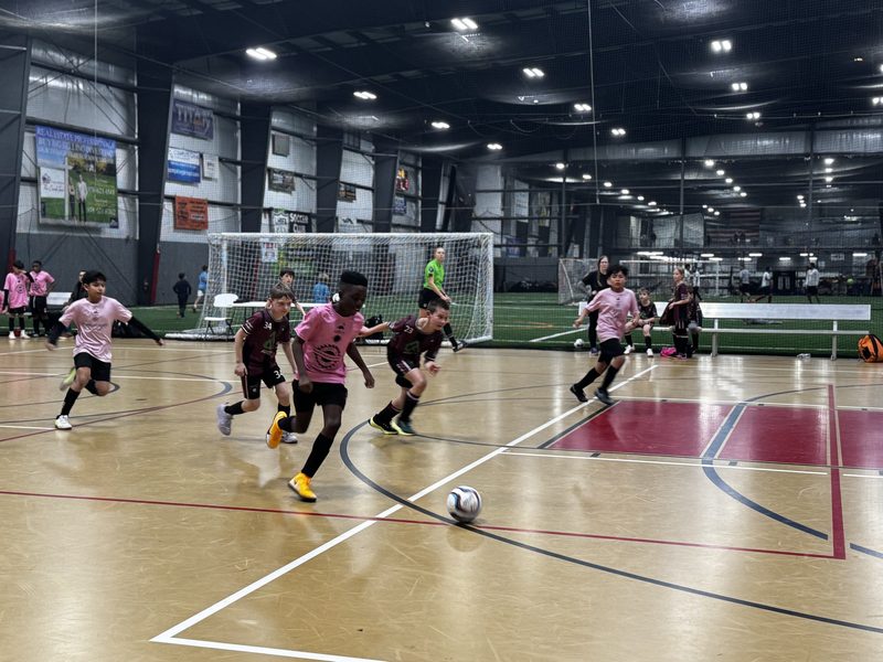 Players in pink and maroon jerseys sprinting after the ball on an indoor futsal court