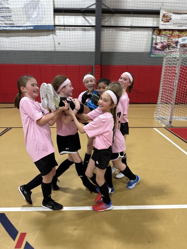 A girls team in pink jerseys celebrating together on an indoor court, lifting a teammate